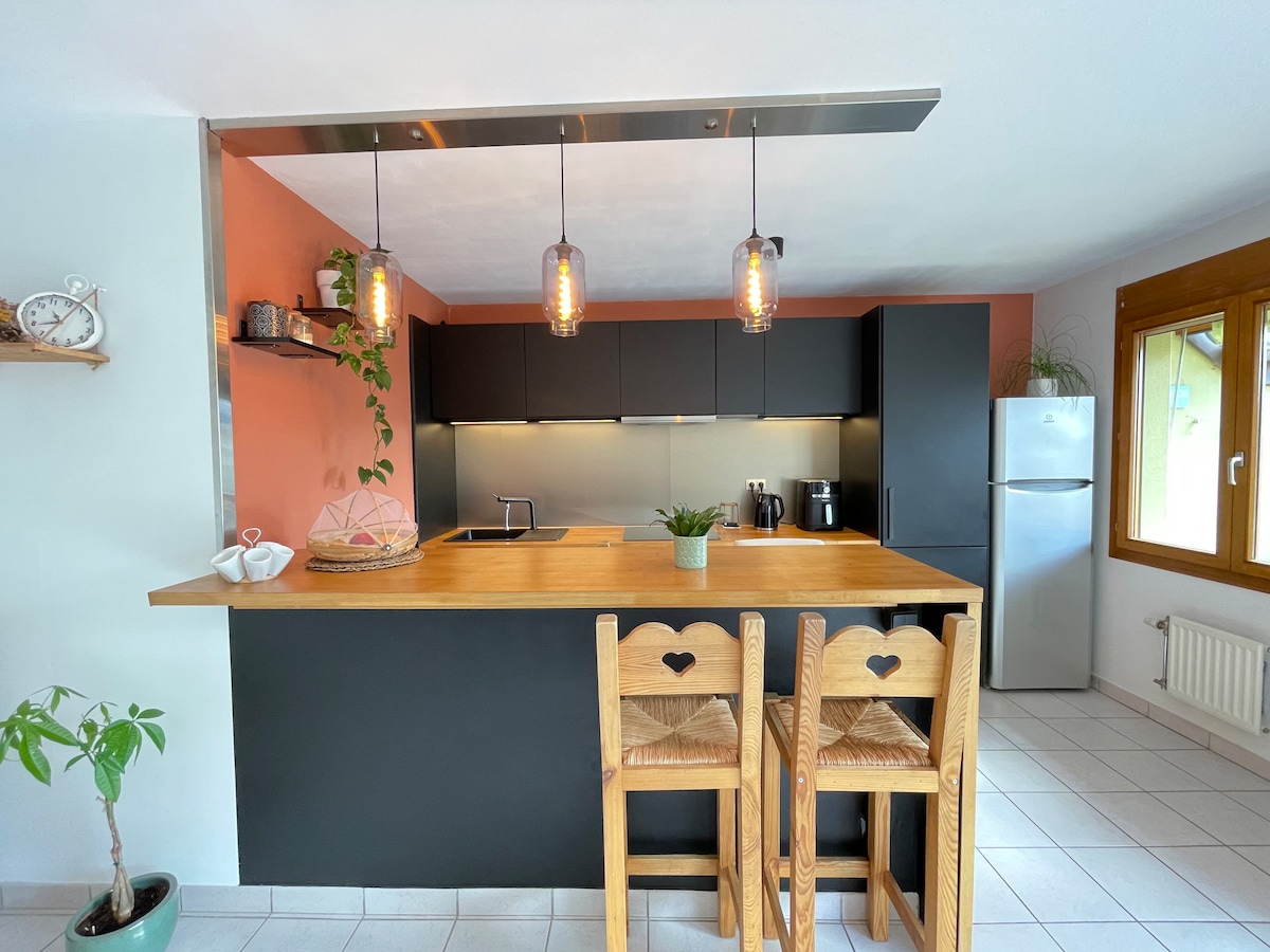 A modern kitchen features a dark cabinetry highlighted by warm pendant lighting. A wooden countertop with two heart-shaped chairs is seen, accompanied by a small potted plant. Stainless steel appliances, including a refrigerator, are positioned against the wall beneath a large window.