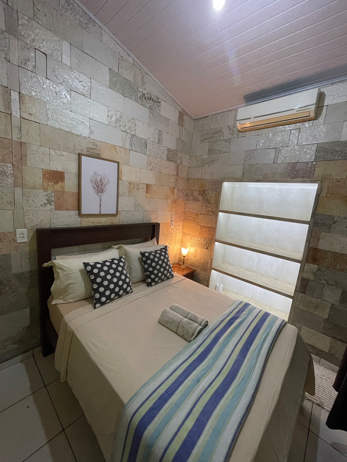 A cozy bedroom features textured stone walls, a spacious bed adorned with neutral linens, and decorative pillows. Soft lighting is provided by a bedside lamp, while a wooden headboard adds warmth. An air conditioning unit is mounted on the wall for comfort.