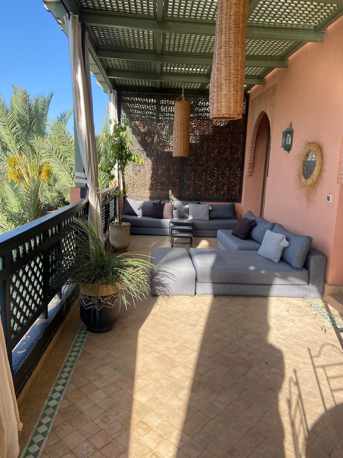 A spacious terrace is equipped with a large grey sectional seating area, adorned with decorative pillows. Natural light fills the area, highlighting the textured walls and green foliage, while wicker pendant lamps hang from the ceiling, creating a calm and inviting environment.