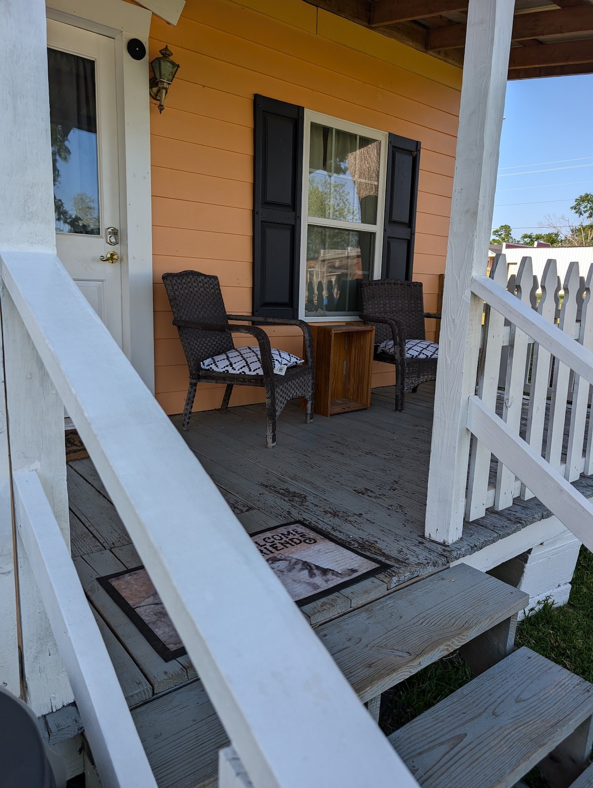 A welcoming porch features two wicker chairs positioned on a wood deck, accompanied by a small table. A doormat is placed at the entrance, and the exterior wall is painted in a soft orange tone, contrasted by dark shutters and a white railing.