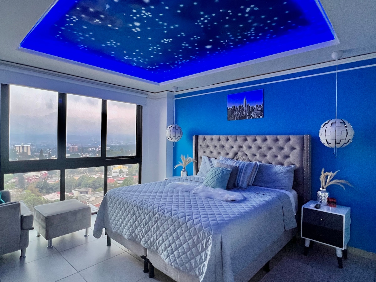 A spacious bedroom is enhanced by a starry ceiling feature and vibrant blue walls. A comfortable bed is dressed with cozy linens, complemented by decorative pillows. Large windows frame city views, while two stylish light fixtures add a modern touch to the space.