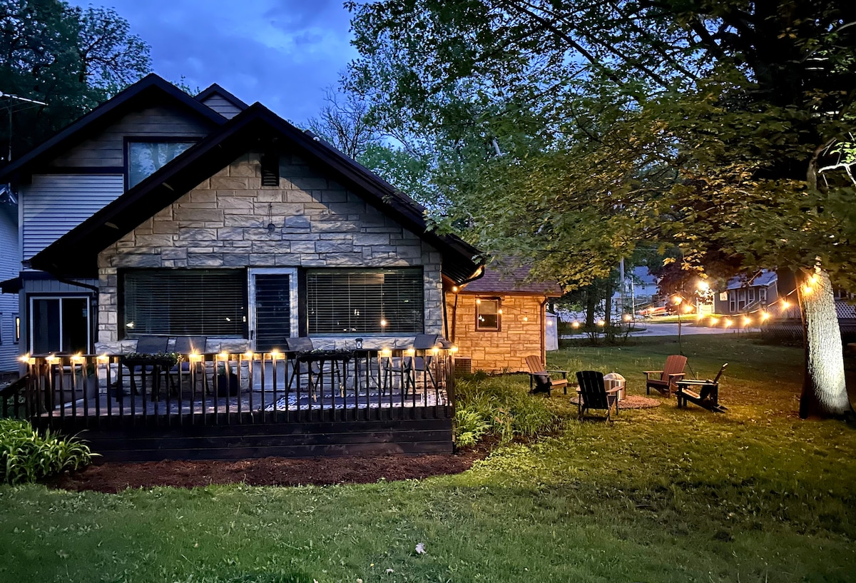 An inviting exterior view of the home showcases a large deck adorned with string lights, creating a warm ambiance. The grassy lawn is highlighted by Adirondack chairs, inviting relaxation. Surrounding trees provide shade, while the house's stone facade adds character against the evening sky.