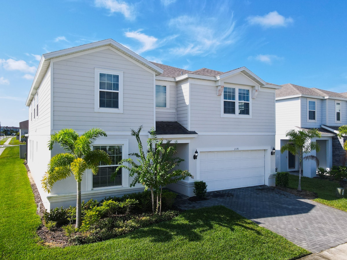 A spacious two-story mansion is depicted, featuring a well-manicured lawn and landscaping. The exterior showcases light-colored siding with multiple windows, while a paved driveway leads to a garage. The home is framed by palm trees, set under a clear blue sky.