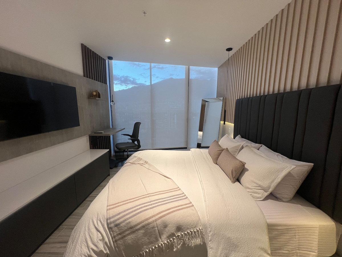 A modern bedroom features a spacious bed with soft bedding and decorative pillows. A large window offers views of the city, while a work desk and a sleek television are visible on the wall. Soft lighting highlights the textured wall design.