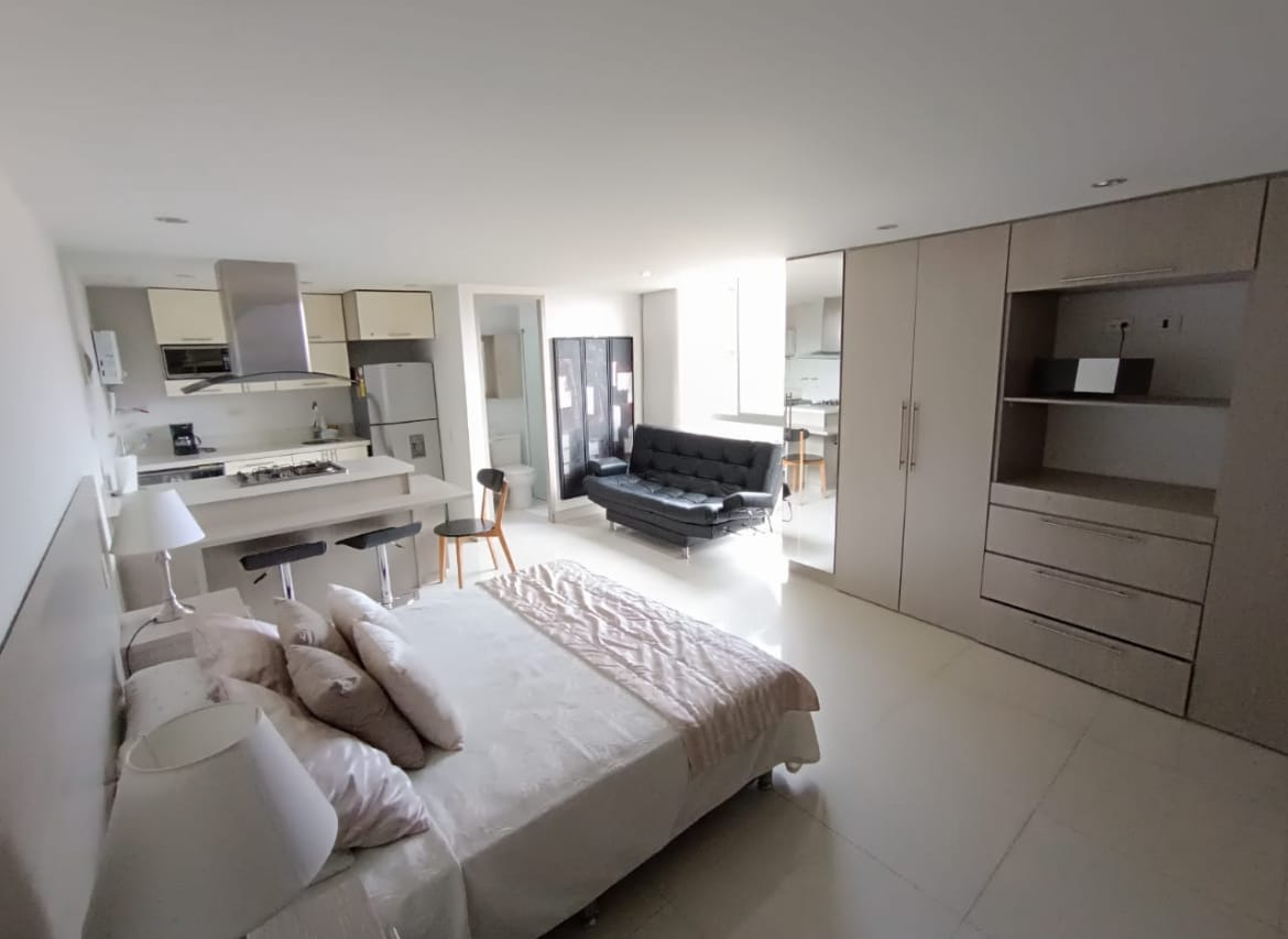 A spacious studio apartment is revealed, featuring a comfortable sleeping area with a light-colored bed and bedside lamps. An adjacent seating space includes a dark sofa bed and a stool, while a modern kitchen showcases sleek cabinetry and appliances.