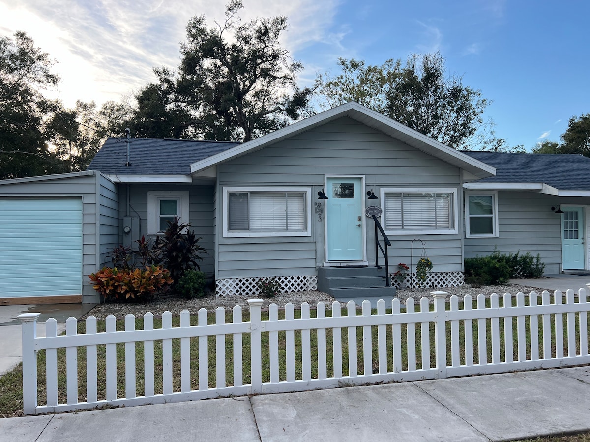 A charming cottage, painted in soft blue, features a welcoming front porch and a white picket fence. Lush green plants are arranged along the front path, and a light-colored garage door complements the exterior. Windows with white blinds allow natural light to fill the home.