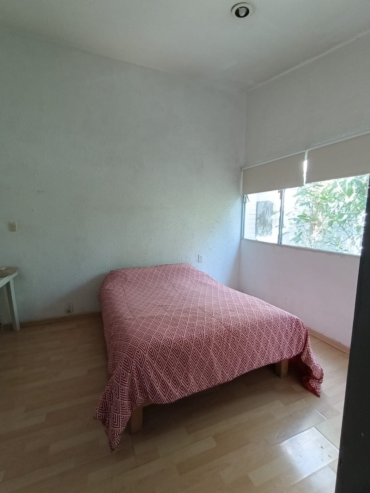 A simple sleeping area features a bed covered with a patterned red blanket. Natural light enters through a window, which is adorned with light-filtering shades. The room has light wood flooring and a neutral wall color.