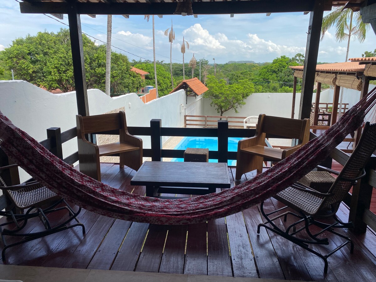 A shaded outdoor seating area features a textured hammock suspended between wooden supports, with a view of a turquoise pool. Wooden chairs and a small table are arranged on a rich wooden deck, framed by greenery and a clear blue sky above.