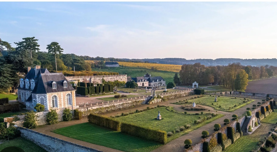 An expansive landscape features manicured gardens with neatly trimmed hedges and pathways, framed by vibrant greenery. Several elegant structures, including a grand house, are visible, surrounded by open fields and trees in the background, reflecting the tranquil charm of the countryside.