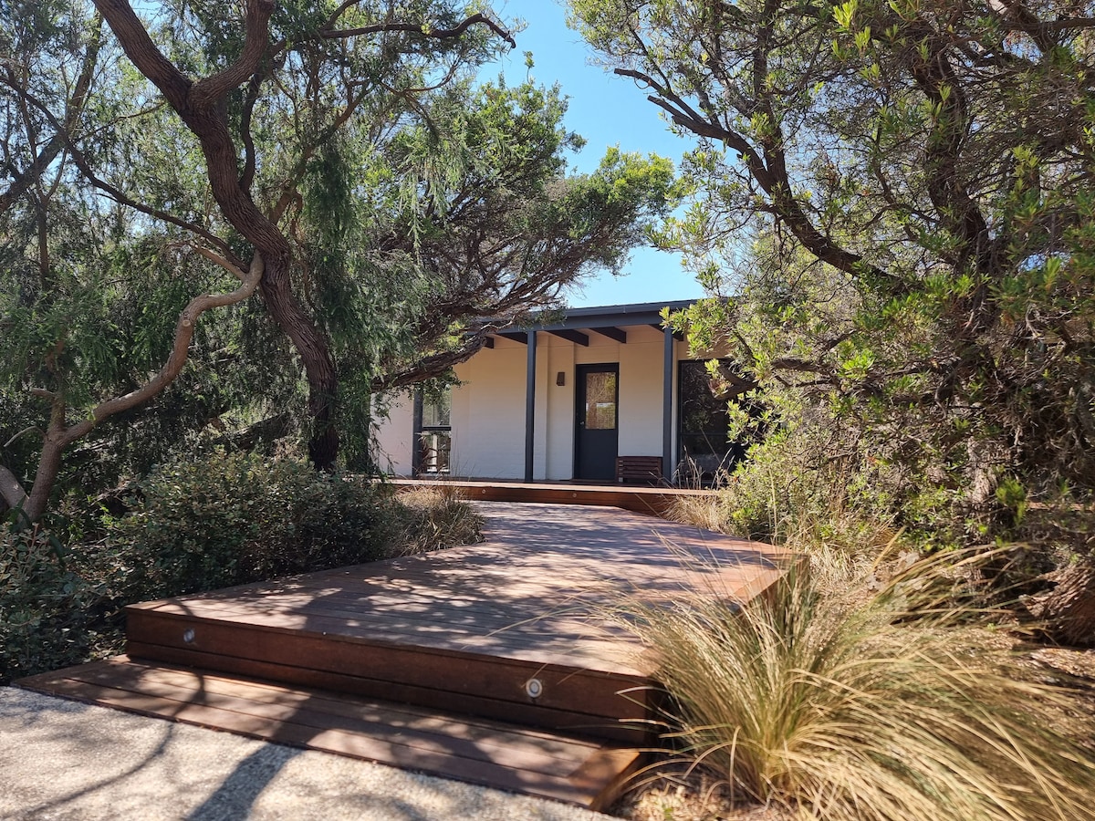 The entrance path leading to the coastal shack is framed by native vegetation, showcasing a blend of textured wood decking and natural landscaping. The facade of the house is partially visible, providing an inviting glimpse into the tranquil setting.