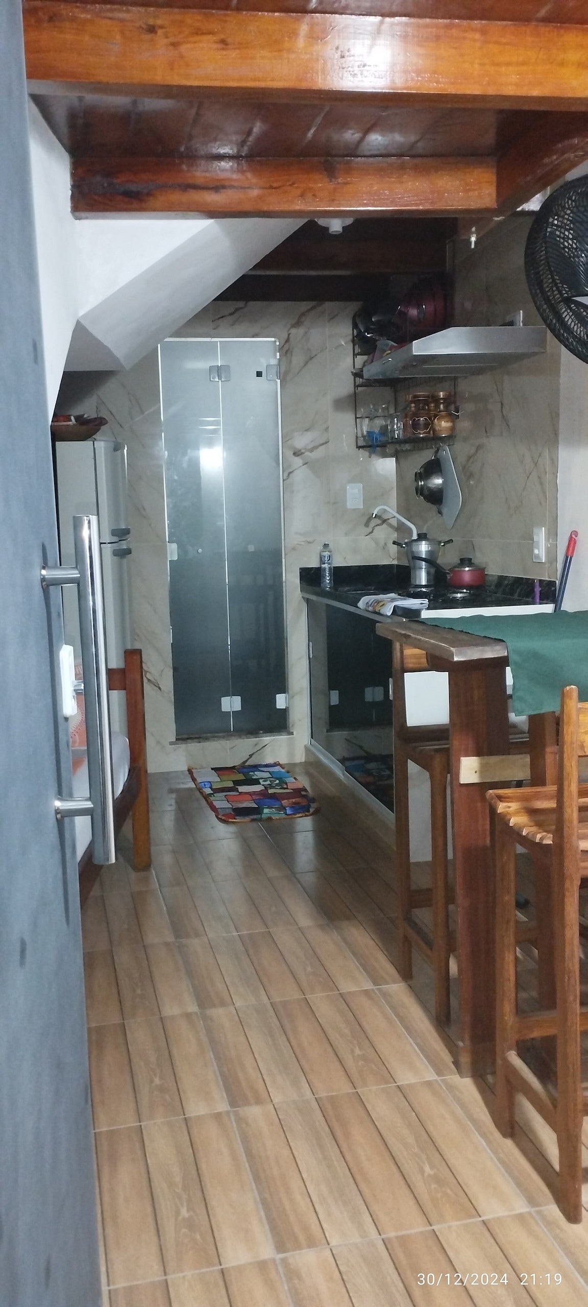 A compact kitchen area features a black countertop with essential cooking appliances, including a stove and refrigerator. Wooden stools are positioned at a small bar. Bright tiles and wood accents create an inviting ambience, while natural light illuminates the space.