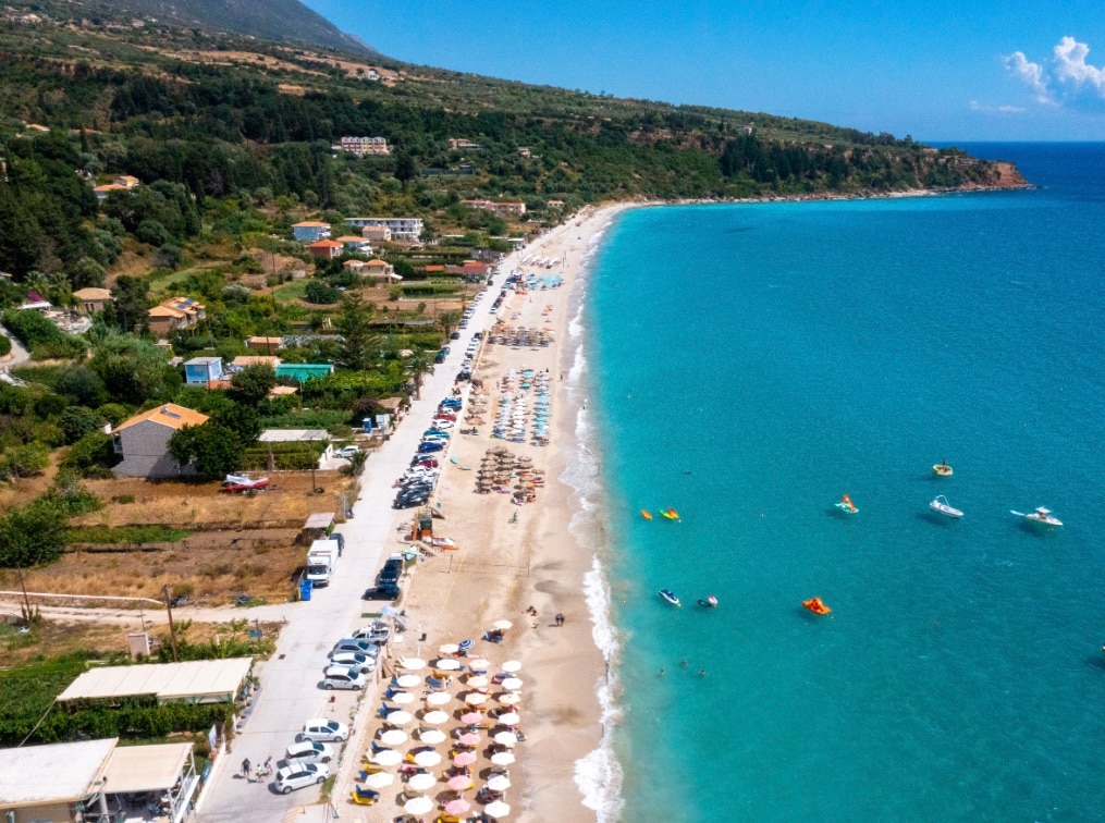 A long stretch of sandy beach is seen alongside a vibrant blue sea, dotted with boats. Beachgoers can be spotted under sun umbrellas on the shore. Villas and green landscapes are visible in the background, contributing to the coastal charm of the location.