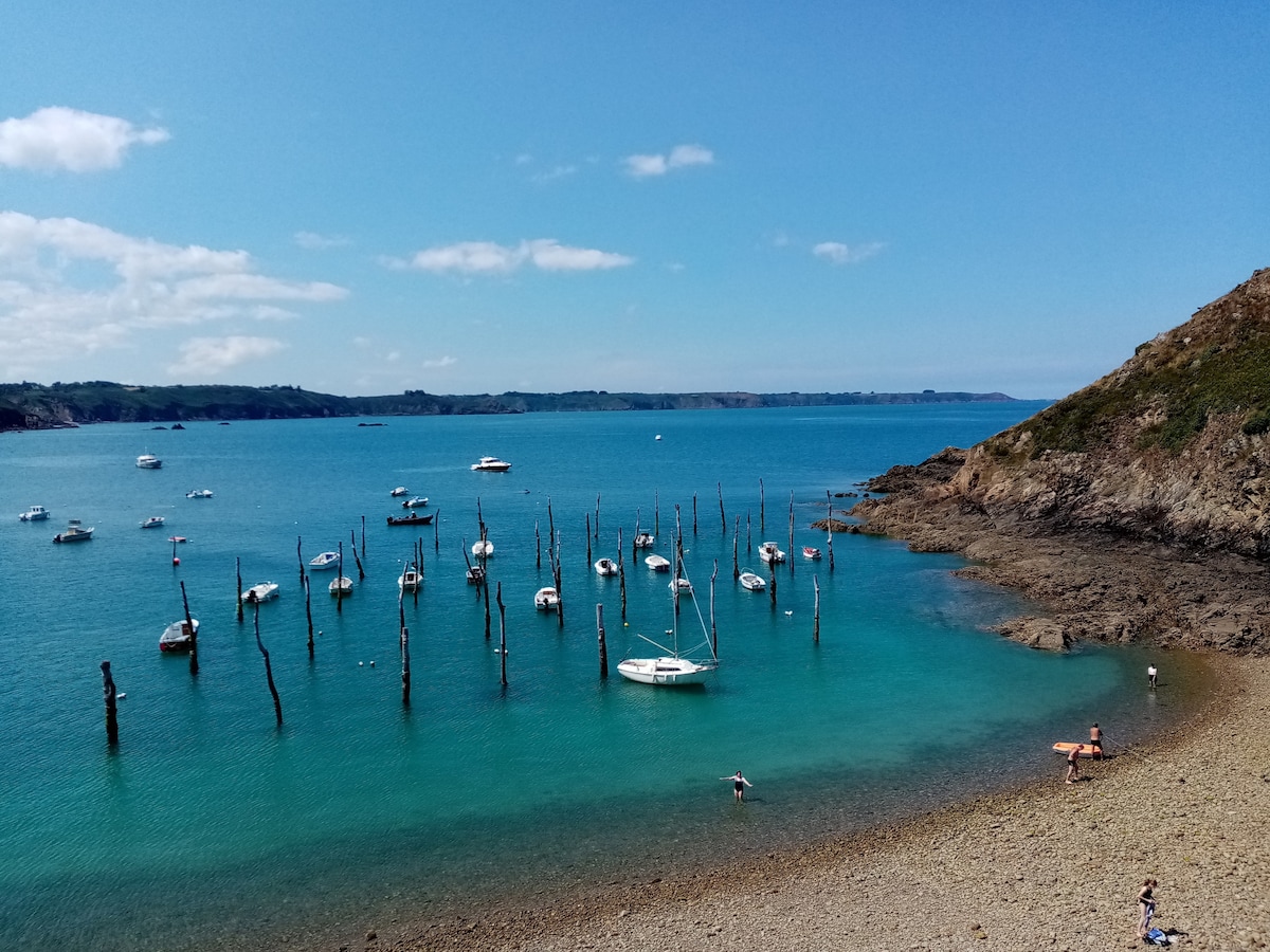 A serene coastal landscape captures a calm blue sea dotted with several boats anchored near a rocky shore. Tall, weathered posts rise from the water, adding a unique element to the scene. A sandy beach with a few people enjoying the sun enhances the tranquil atmosphere.