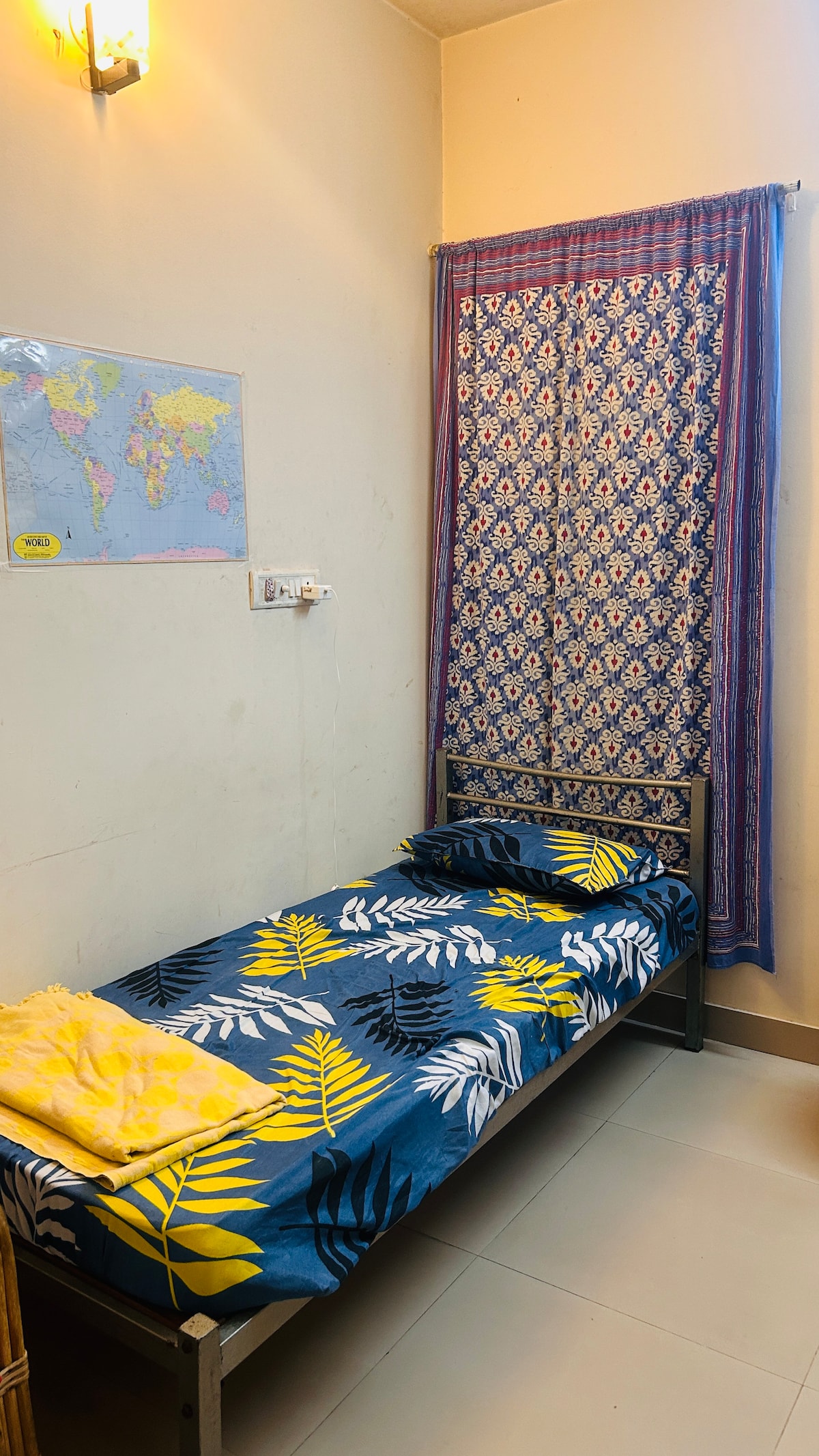 A small bedroom features a bed dressed in a colorful, tropical-patterned cover. A light yellow towel is neatly placed on the bed. A decorative curtain with intricate patterns drapes over the window, allowing natural light to brighten the room.