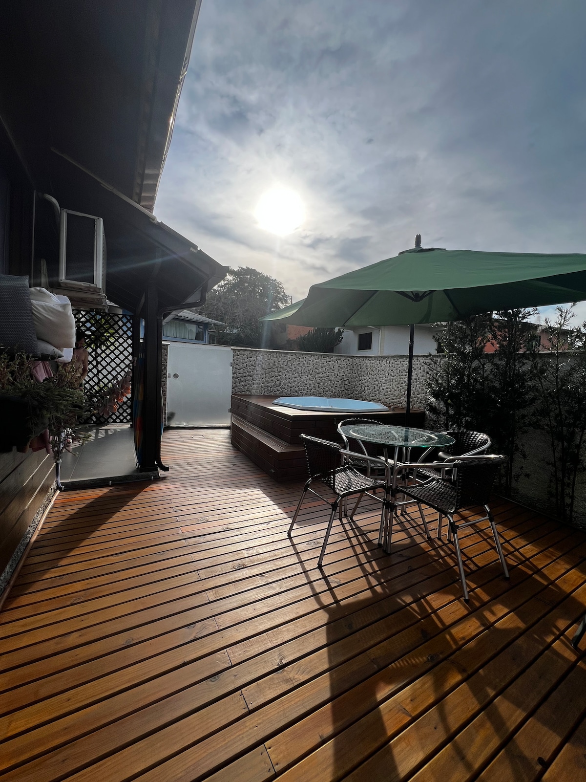 A spacious outdoor terrace features a wooden deck, with sunlight streaming down. A small hot tub is visible alongside a round table and four metal chairs, providing a comfortable space for relaxation. An umbrella offers shade from the sun, enhancing the inviting atmosphere.