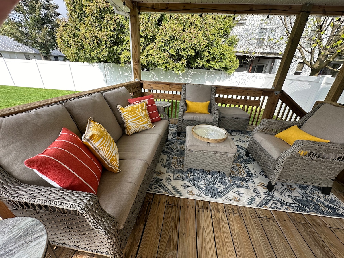 A covered outdoor seating area features a plush sectional sofa with yellow and red accent pillows. A simple coffee table sits in the center, positioned on a decorative area rug. Surrounding greenery creates a serene backdrop, enhancing the cozy atmosphere.