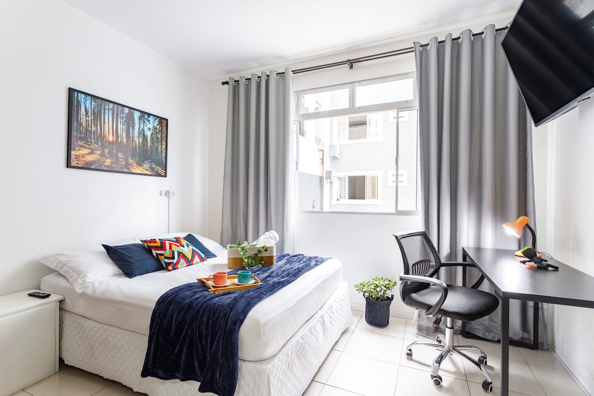 A bright bedroom is furnished with a spacious bed, adorned with colorful cushions and a soft blanket. Natural light enters through large windows, which are framed by gray curtains. A desk with an office chair and a small plant create a functional work area.
