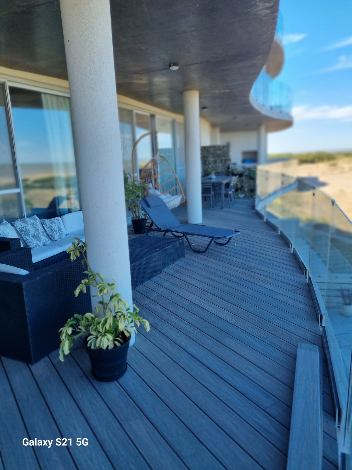 A spacious balcony is shown with wooden decking and a clear glass railing. Comfortable seating arrangements, including lounge chairs and a wicker chair, overlook scenic views. A potted plant adds a touch of greenery, enhancing the outdoor space.