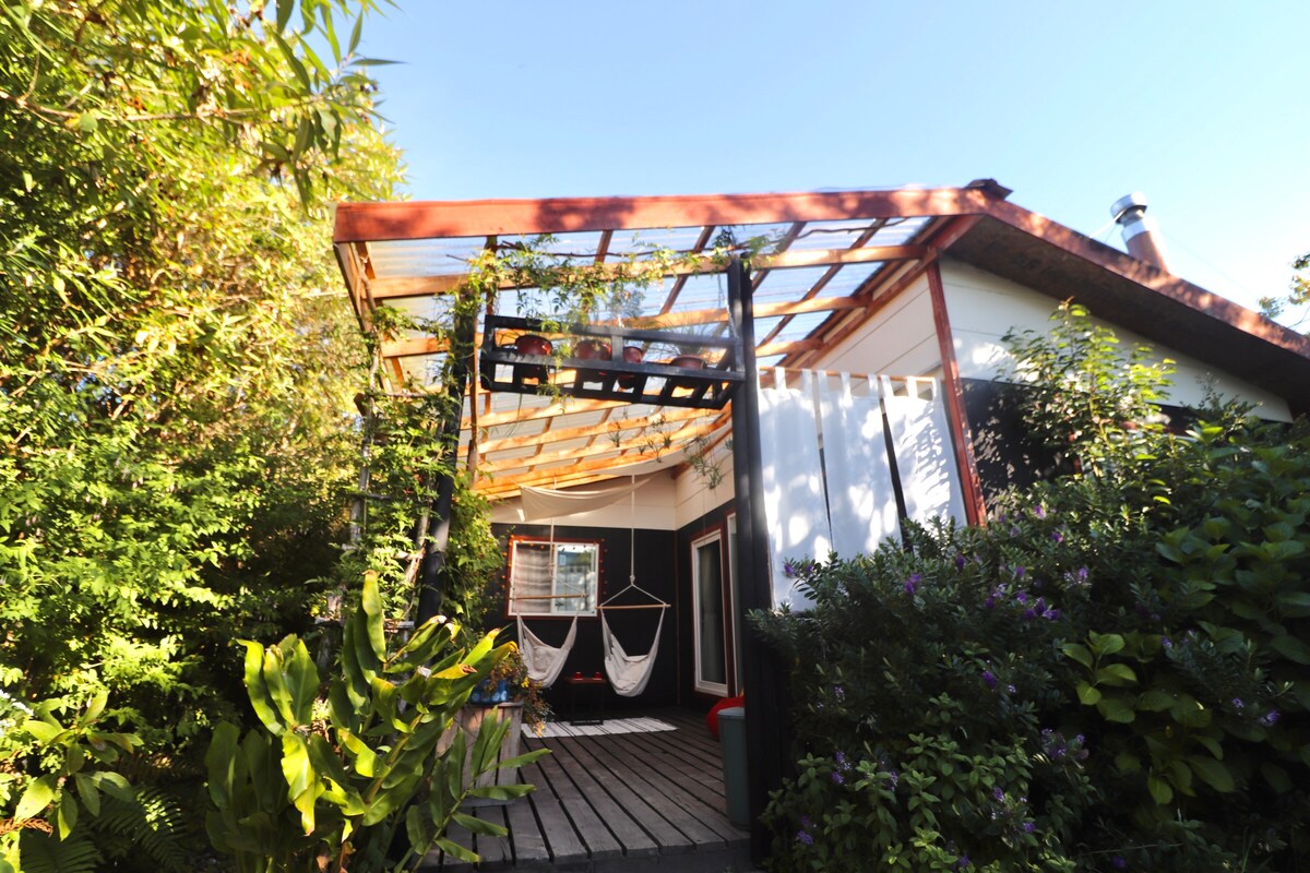 The exterior of the cabin features a deck surrounded by lush greenery. Two hammocks are suspended under a slatted roof, allowing natural light to filter through. A vibrant garden and flowering plants contribute to a serene outdoor space.