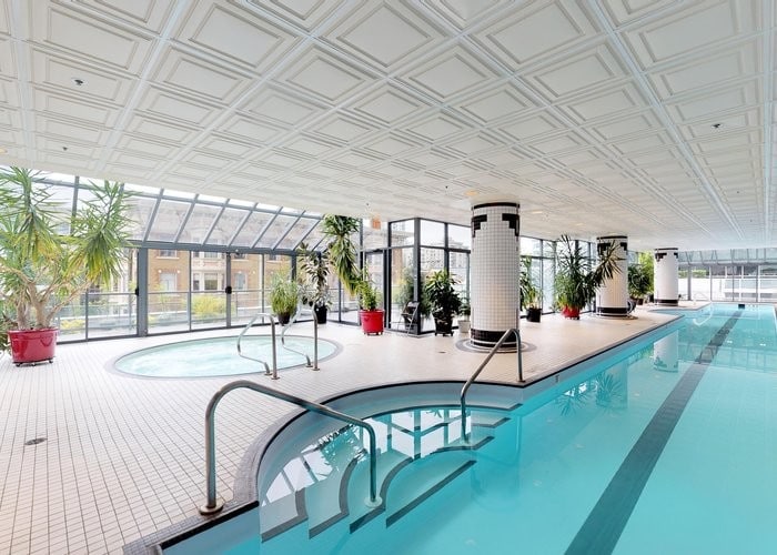 An indoor swimming pool area is highlighted by large windows, allowing natural light to illuminate the space. Potted plants are placed around the room, adding greenery. A separate hot tub is visible beside the pool, with tiled surfaces providing a clean and spacious environment for relaxation.
