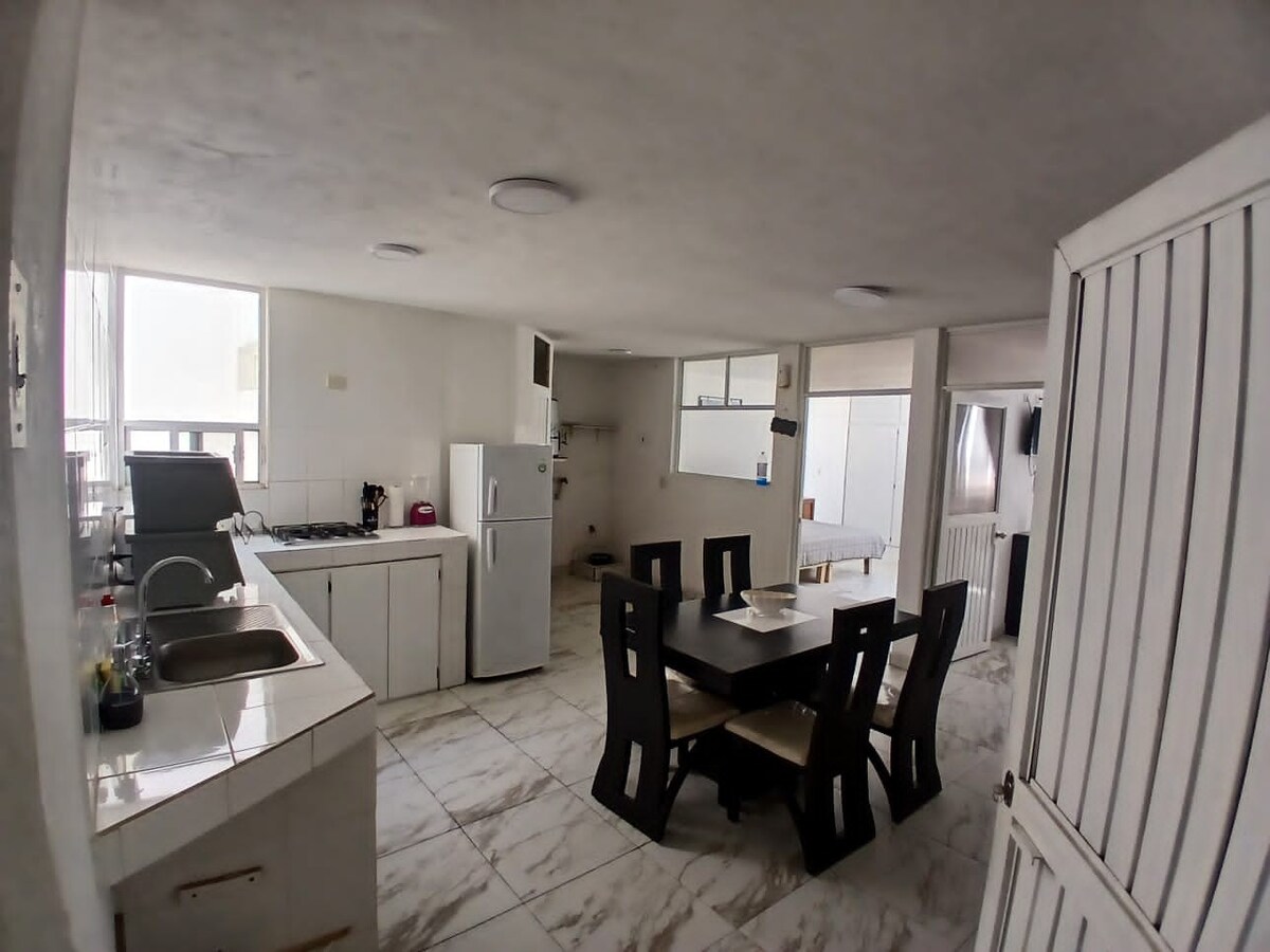 A spacious kitchen area features a dining table with six chairs, complemented by a refrigerator, stovetop, and sink. Light floods the room through large windows, and neutral-colored walls enhance the simplicity of the design. A door leads to the adjoining space.