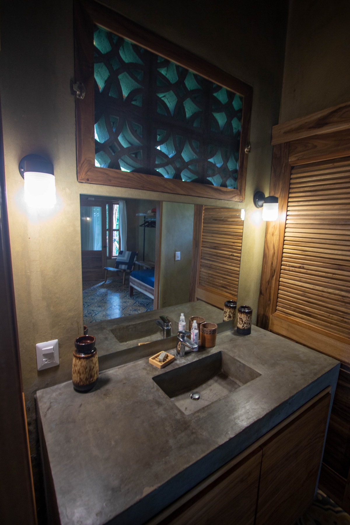 A modern bathroom features a concrete sink atop wooden cabinetry, with decorative elements including a patterned mirror and wall sconces. Natural light is allowed to enter through the window, reflecting off the smooth surfaces and enhancing the overall spaciousness.