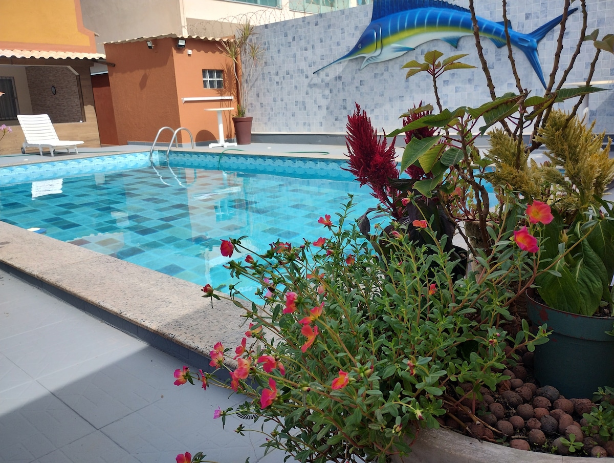 A well-maintained swimming pool reflects the clear blue sky, surrounded by colorful plants and flowers. A decorative mural of a marlin is visible on the wall, adding a touch of artistic charm to the area. Sun loungers are positioned nearby for relaxation.