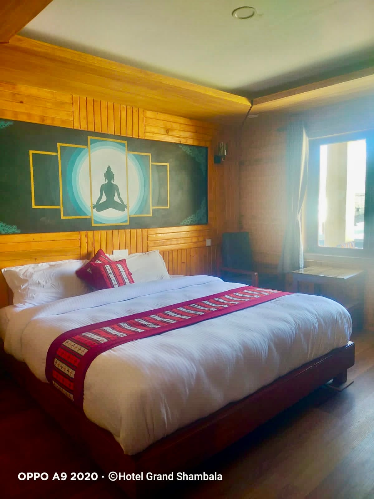 A cozy sleeping space features a spacious bed adorned with crisp linens and a decorative blanket. A serene mural of a Buddha figure is displayed on the wooden wall. Natural light enters through a window, enhancing the warm tones of the room.