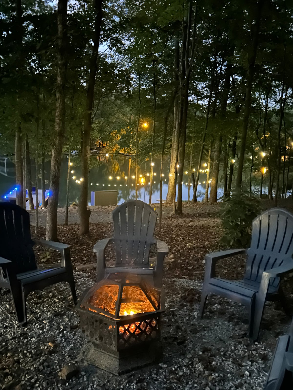 A cozy fire pit sits surrounded by four Adirondack chairs, nestled among trees. Soft string lights illuminate the area, casting a warm glow over the gravel. The lake is visible through the trees, enhancing the serene environment.