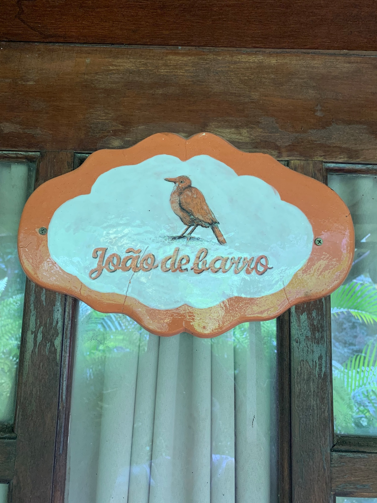 A decorative sign hangs above a wooden door, featuring an illustration of a bird and the name 'João de barro' in a elegant script. Soft natural light filters through the nearby glass panes, highlighting the craftsmanship of the sign and the surrounding greenery.