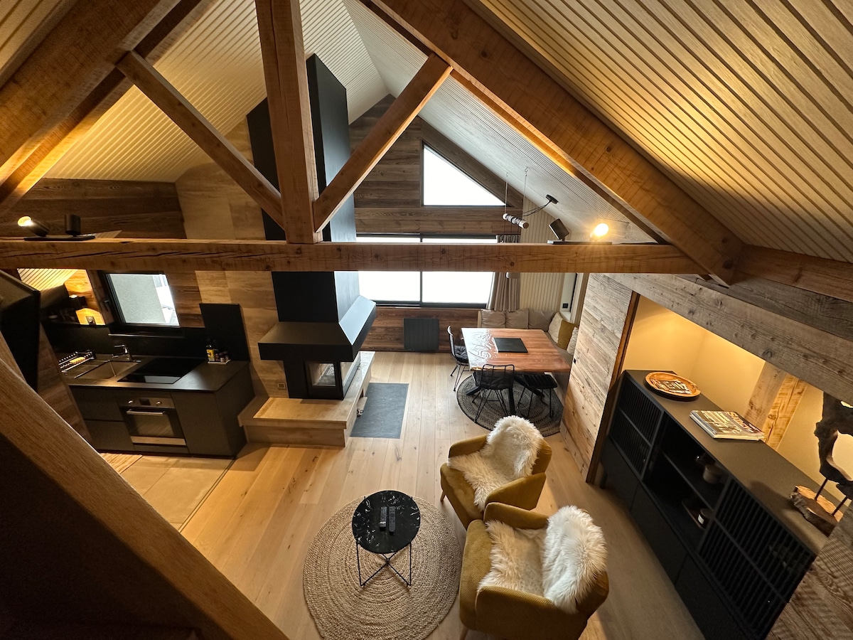 An open-plan living area features a high ceiling with exposed wooden beams. A cozy seating arrangement includes two armchairs with soft textures, positioned near a coffee table. The modern kitchen is visible, alongside a stylish dining table. A fireplace provides warmth, enhancing the inviting ambiance.