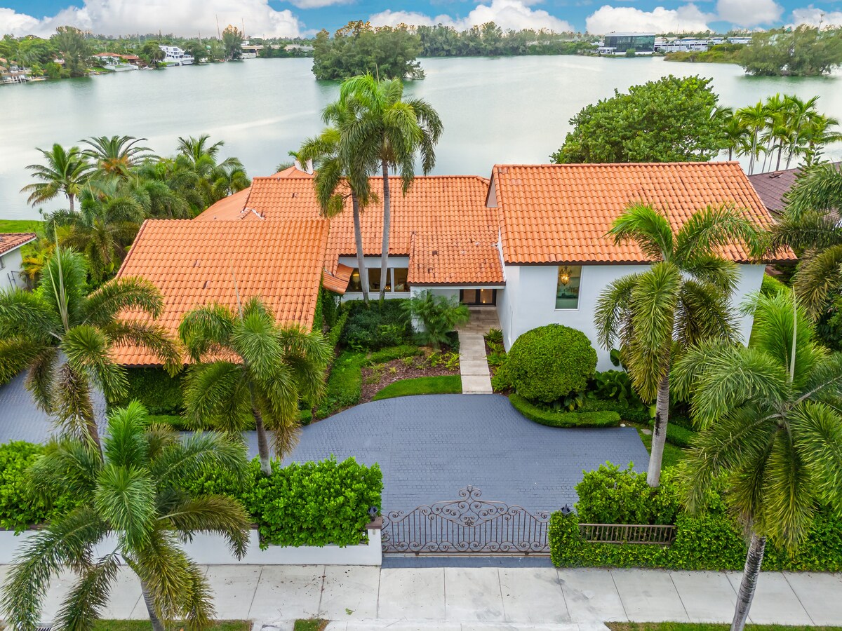 A spacious home is presented from an aerial view, featuring a terracotta tiled roof and palm trees lining the entrance. The manicured lawn leads to a decorative gate that opens to the path toward the house, complemented by lush greenery and views of the lake.