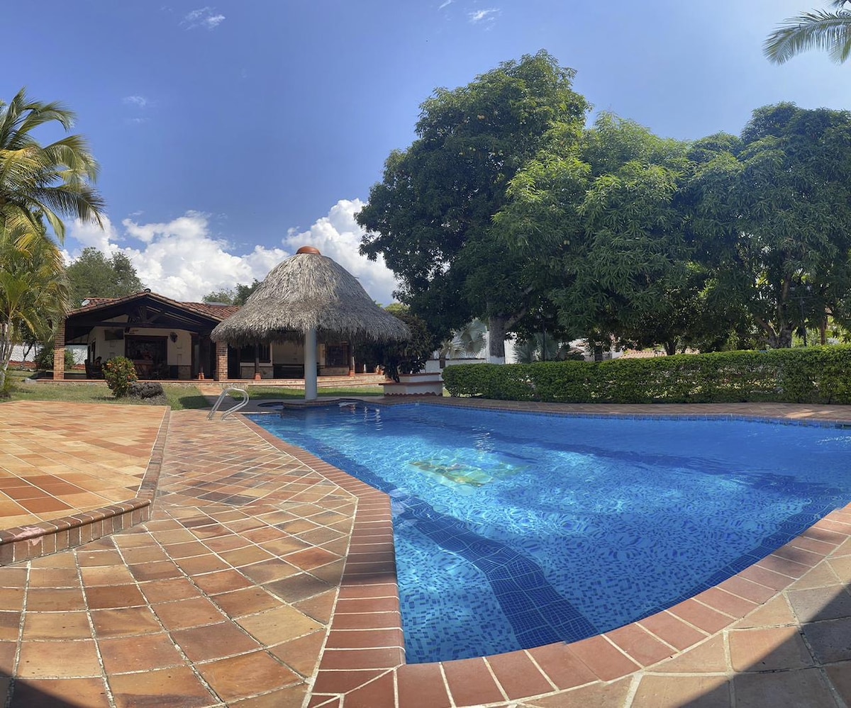 The image shows a spacious pool area surrounded by tropical landscaping. A thatched-roof kiosk is visible in the background, offering shade near the pool. The water of the pool reflects the clear blue sky and lush greenery, providing a serene outdoor setting.