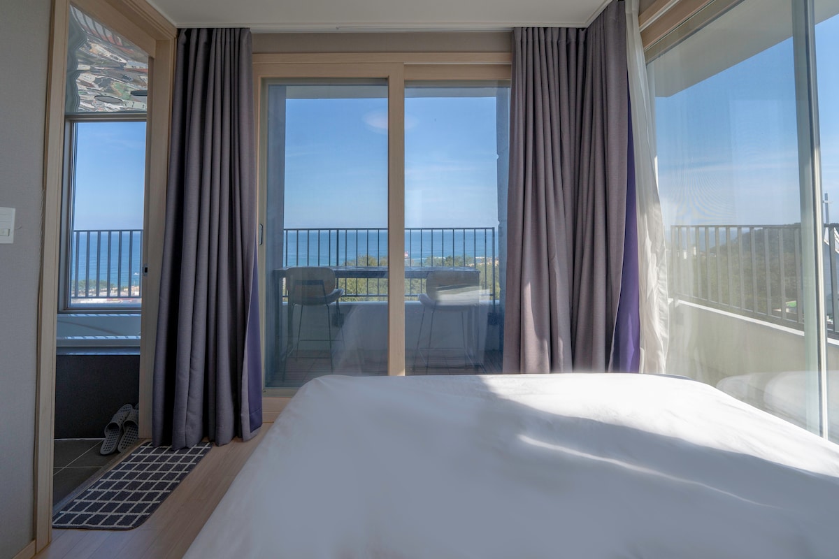A serene bedroom features a spacious bed with crisp white bedding. Large glass doors open to a view of the ocean, allowing natural light to fill the room. Thick curtains frame the windows, and a small table and chairs can be seen on the terrace.