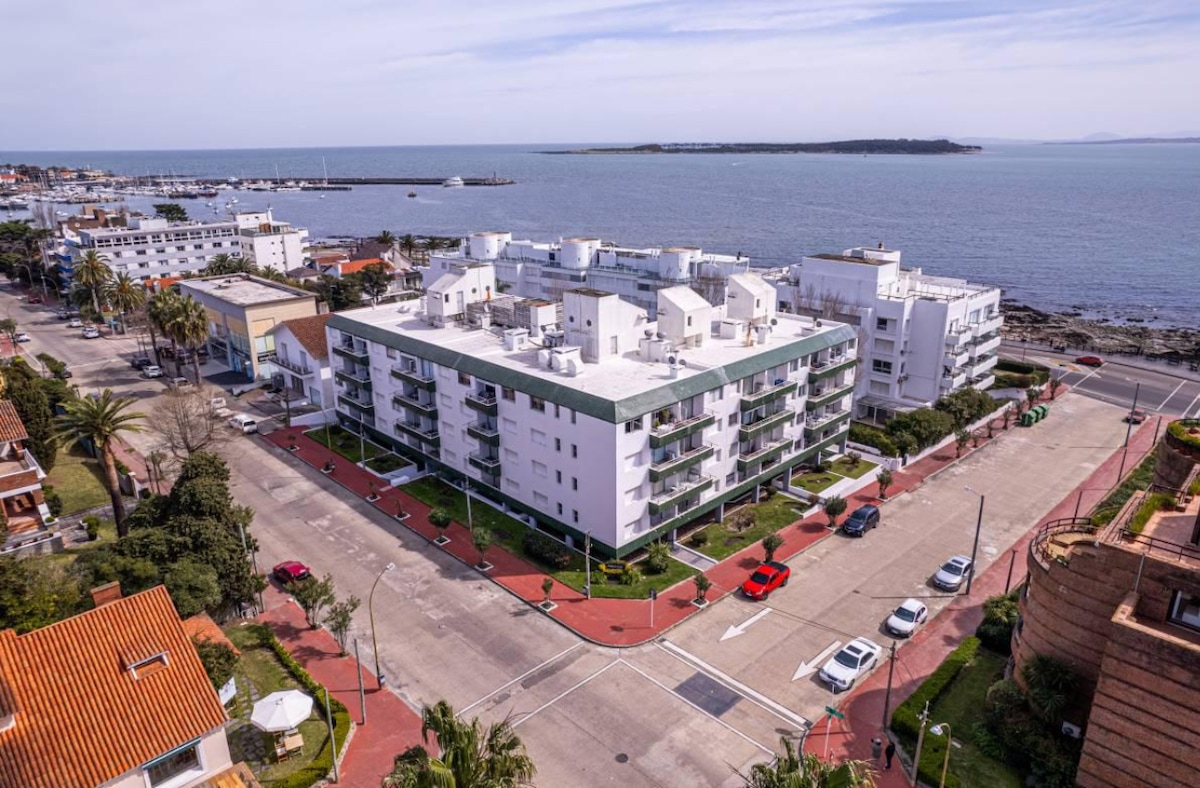 An aerial view captures a modern white building situated at the intersection of two roads, with a marina and ocean visible in the distance. Surrounding greenery and palm trees outline the landscape, while parked cars are seen along the streets.