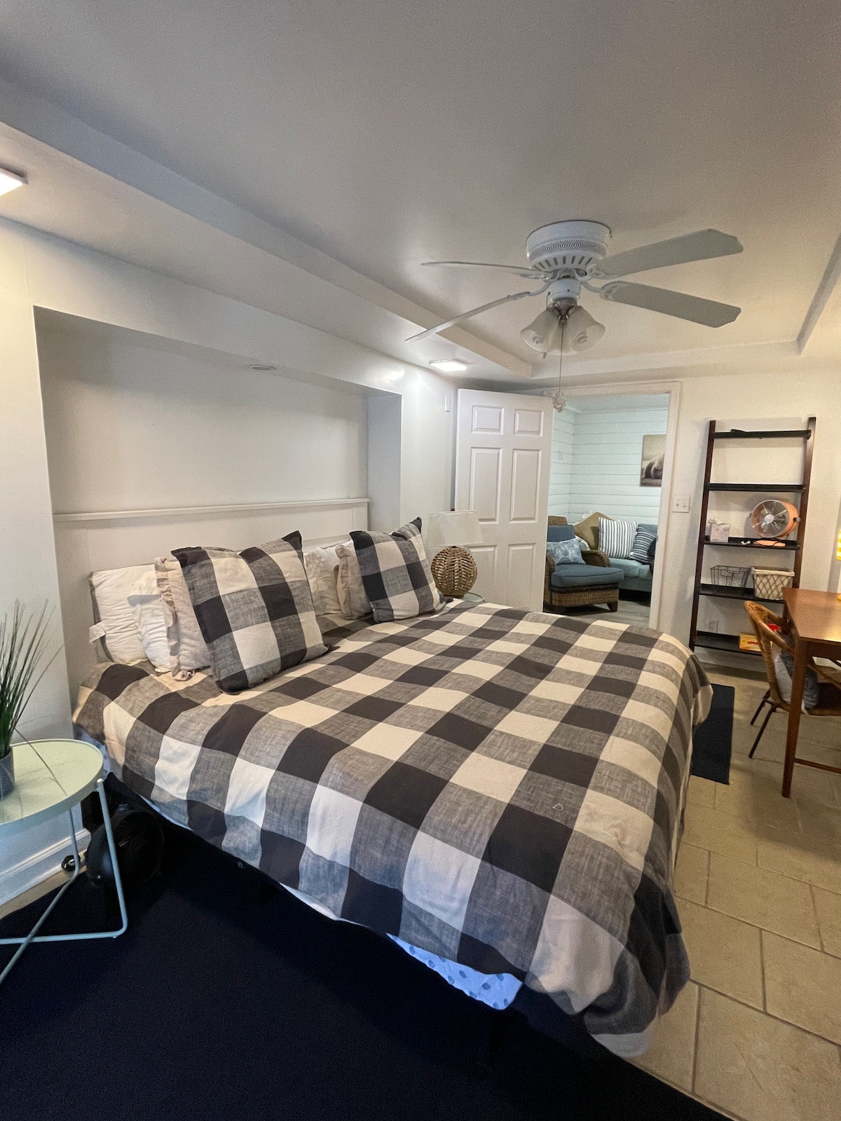 A spacious bedroom features a king bed adorned with checkered bedding and plump pillows. A ceiling fan provides airflow while wood accents and a small plant add touches of nature. A dining table and chair set, along with an open doorway leading to another space, are visible.