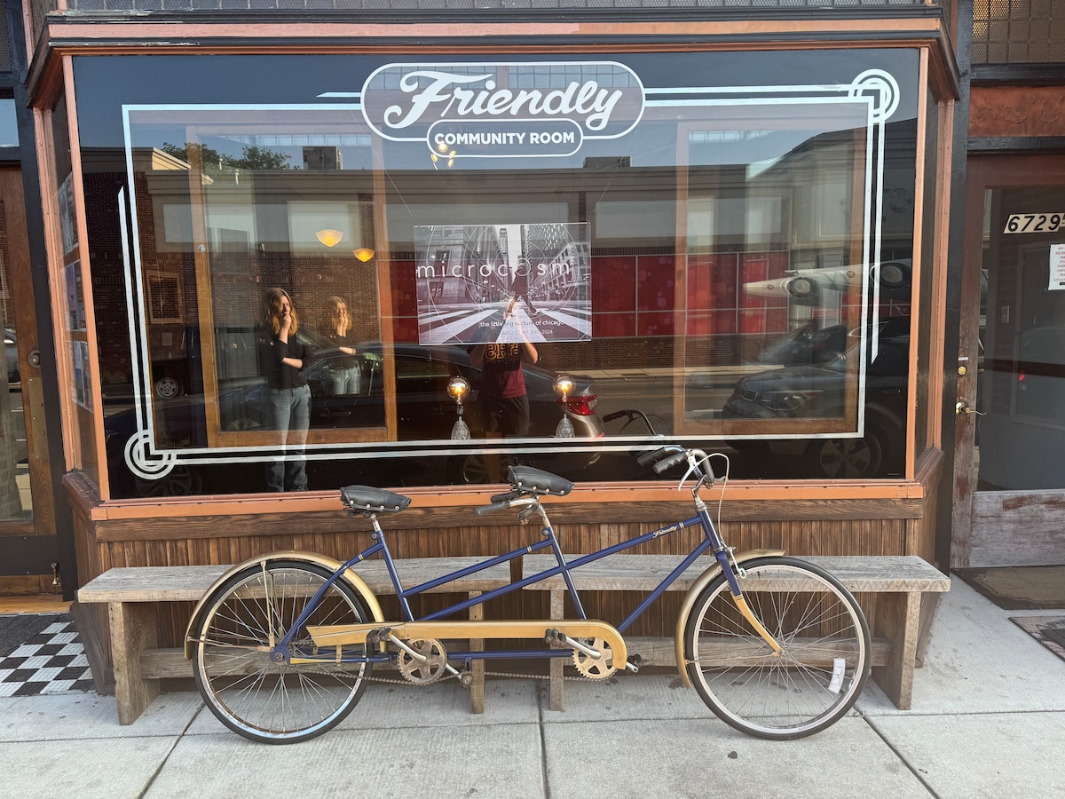 A tandem bicycle is positioned in front of the Friendly Community Room, showcasing a blue frame with yellow accents. The cafe's large window displays the name and a striking piece of art inside, with reflections of passersby visible on the glass.
