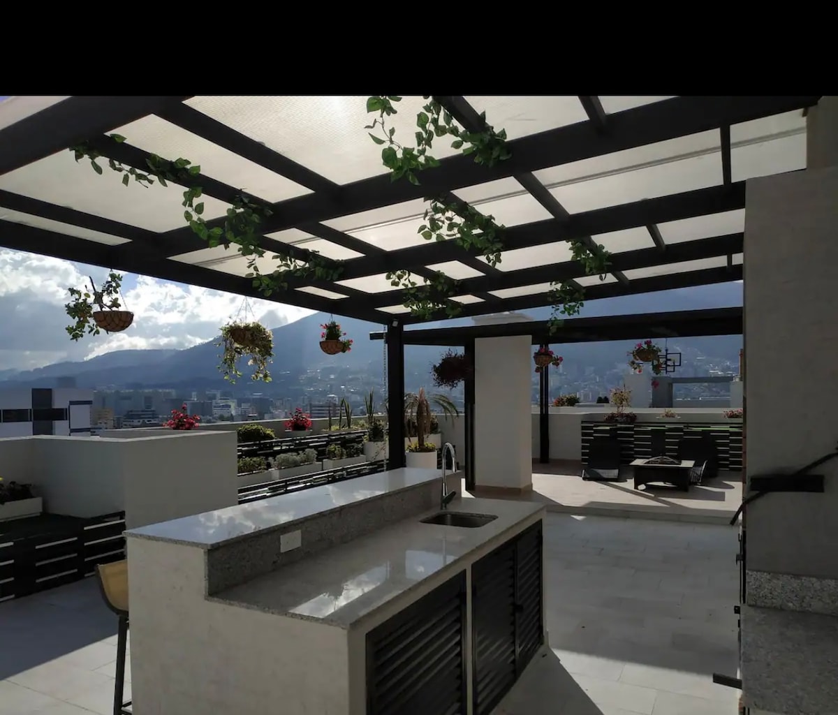 An outdoor terrace is presented with a shaded pergola, adorned with hanging plants. A stone countertop features a sink, while multiple seating areas are arranged for relaxation. Scenic views of the surrounding mountains and cityscape are visible in the background.