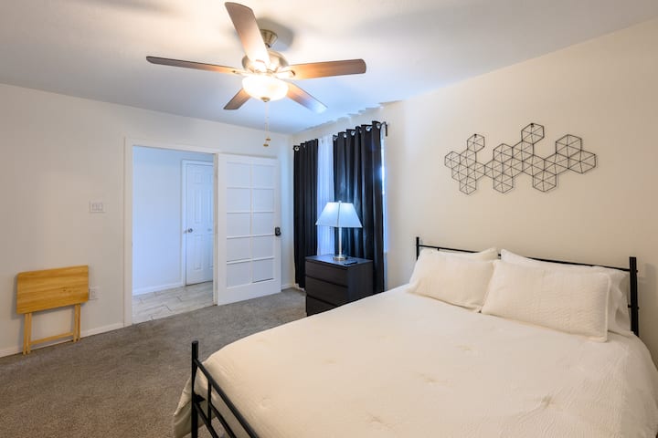 Amplia habitación con colchón tamaño queen, mesita de noche, lámpara, cortinas opacas, ventilador de techo y mesa plegable.