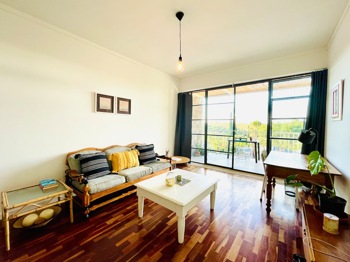 A spacious living area features a comfortable sofa adorned with patterned cushions. A wooden coffee table sits in the center, accompanied by a small side table and a desk beside a large glass sliding door that provides access to the outdoor terrace and surrounding greenery.
