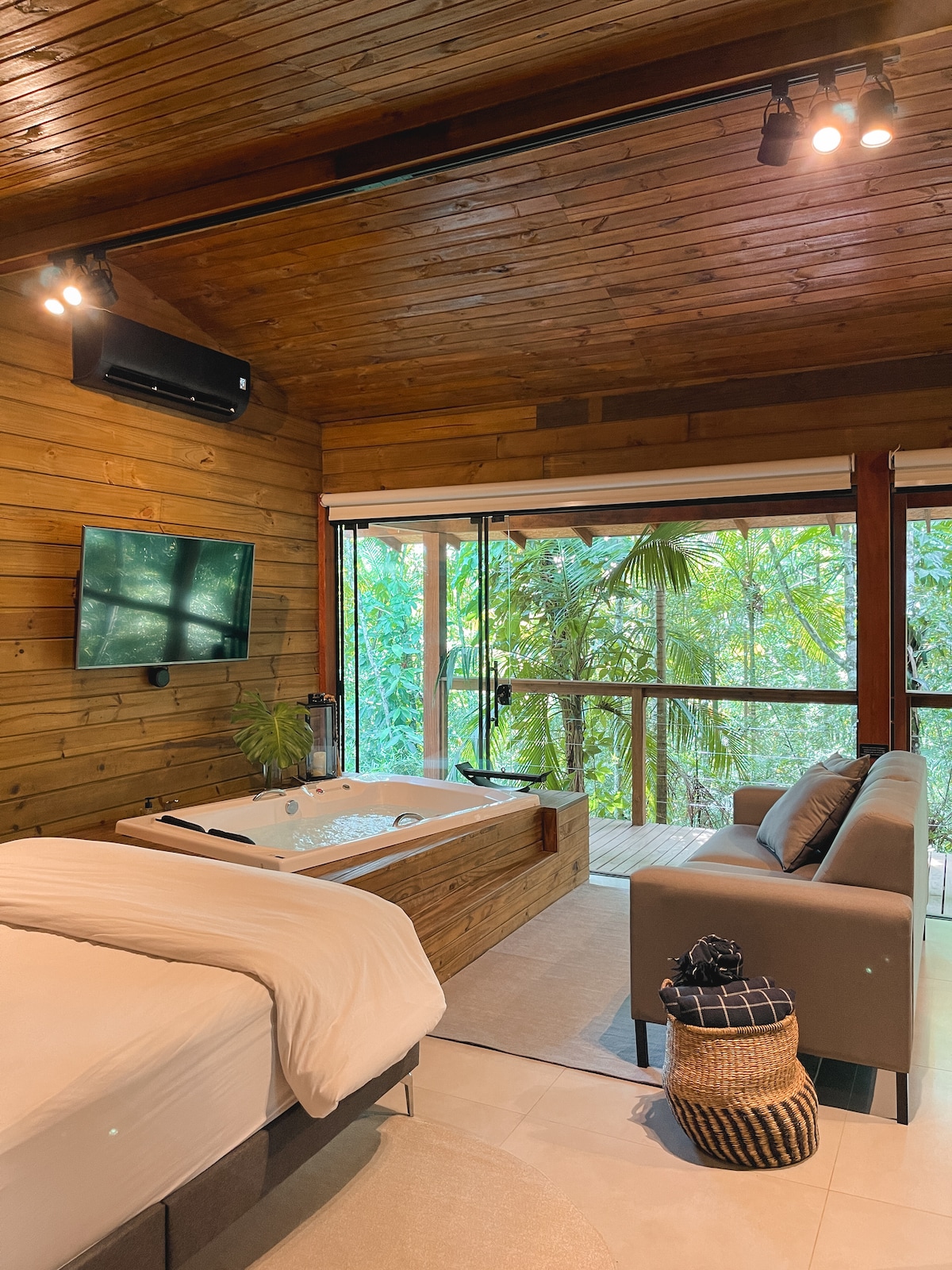 A modern cabin interior features a spacious bed with neutral bedding, a stylish sofa, and a large hot tub set against a backdrop of lush greenery. Natural light filters through expansive glass doors, enhancing the ambiance of relaxation and tranquility.