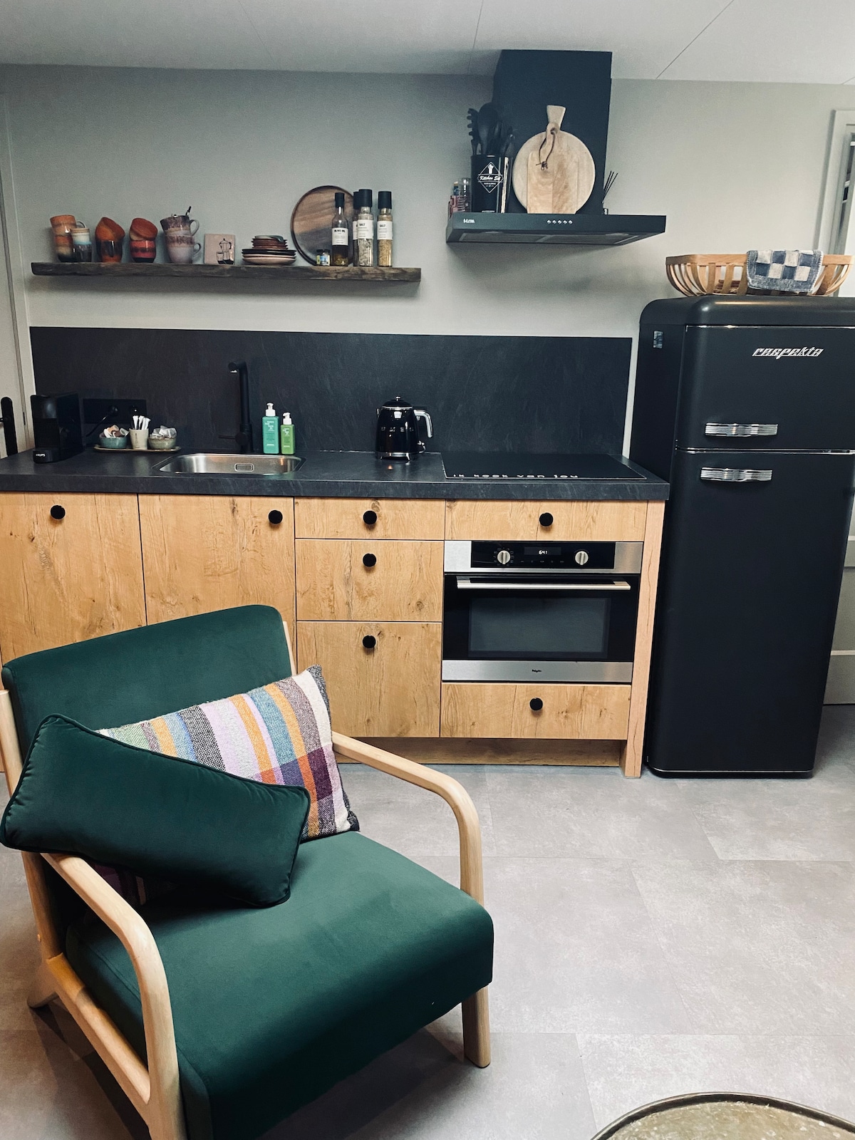A modern kitchen area features wooden cabinetry and a black countertop. A retro-style refrigerator stands beside an oven. A green chair with a colorful pillow is positioned nearby, providing a cozy spot to relax. Shelves display kitchen essentials and decorative items.