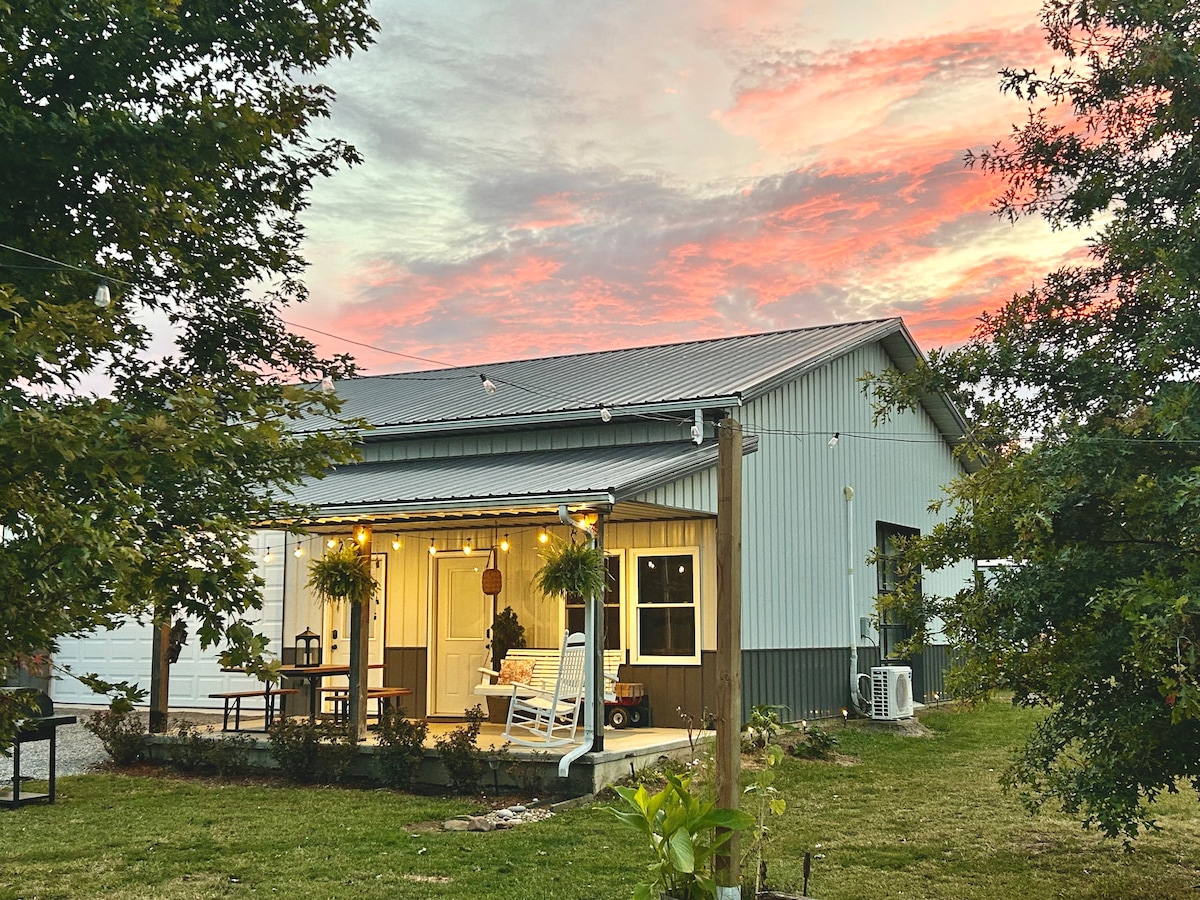 The studio features a covered porch with a swing and picnic table, surrounded by open grass and trees. A calming sunset illuminates the sky, enhancing the inviting appeal of the building, which combines rustic charm with modern elements.