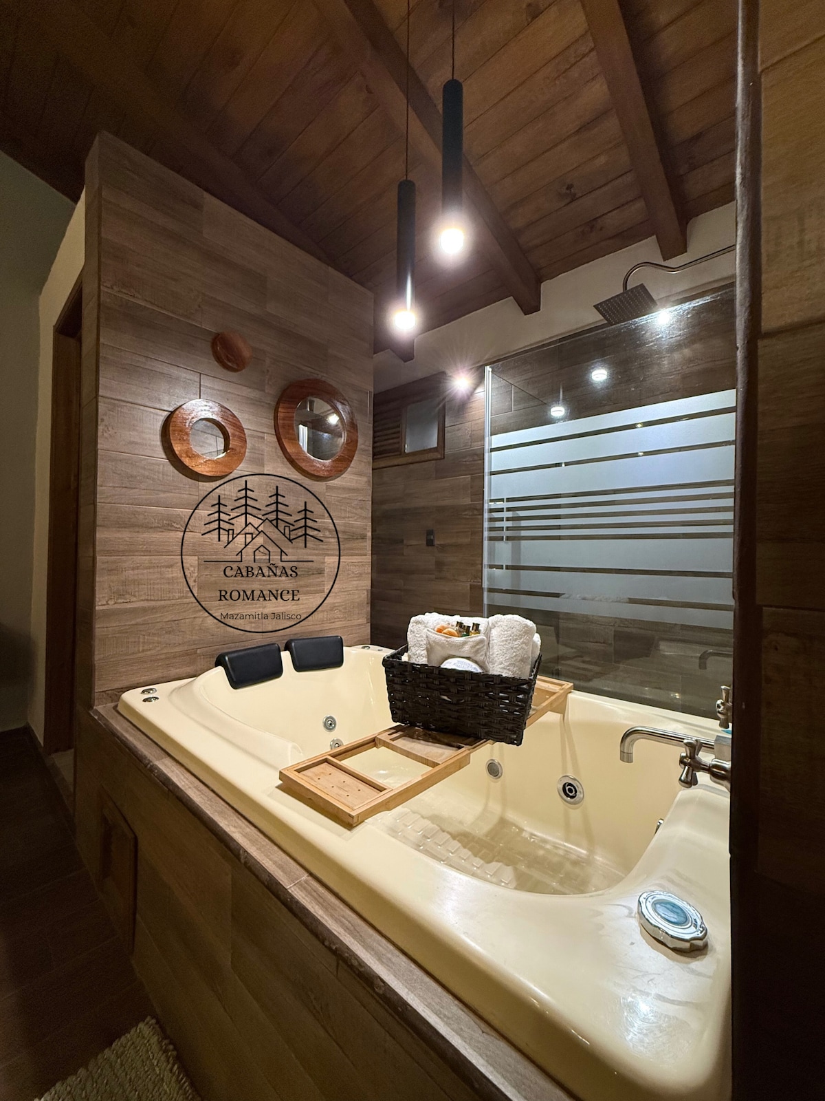 A private bathroom features a spacious jetted tub surrounded by wood accents and natural light. Stylish circular mirrors and modern lighting fixtures enhance the serene ambiance. A wooden tray with rolled towels and a basket adds practical elegance to the inviting space.