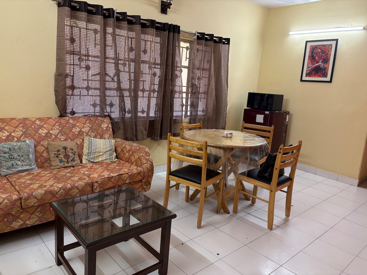 A cozy living space features a patterned couch with decorative cushions, alongside a round dining table with four chairs. A glass coffee table provides a surface for relaxation. Natural light filters through window curtains with a tab design, adding warmth to the environment.