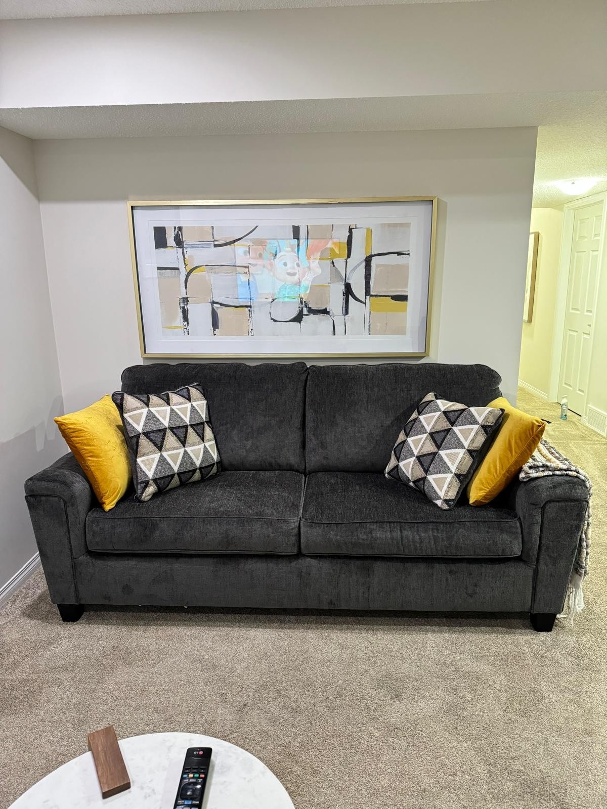 A cozy gray couch with geometric-patterned cushions sits against a light-colored wall. Above the couch is an abstract artwork framed in light wood. A small round coffee table is positioned in front, featuring a remote control and decorative items.