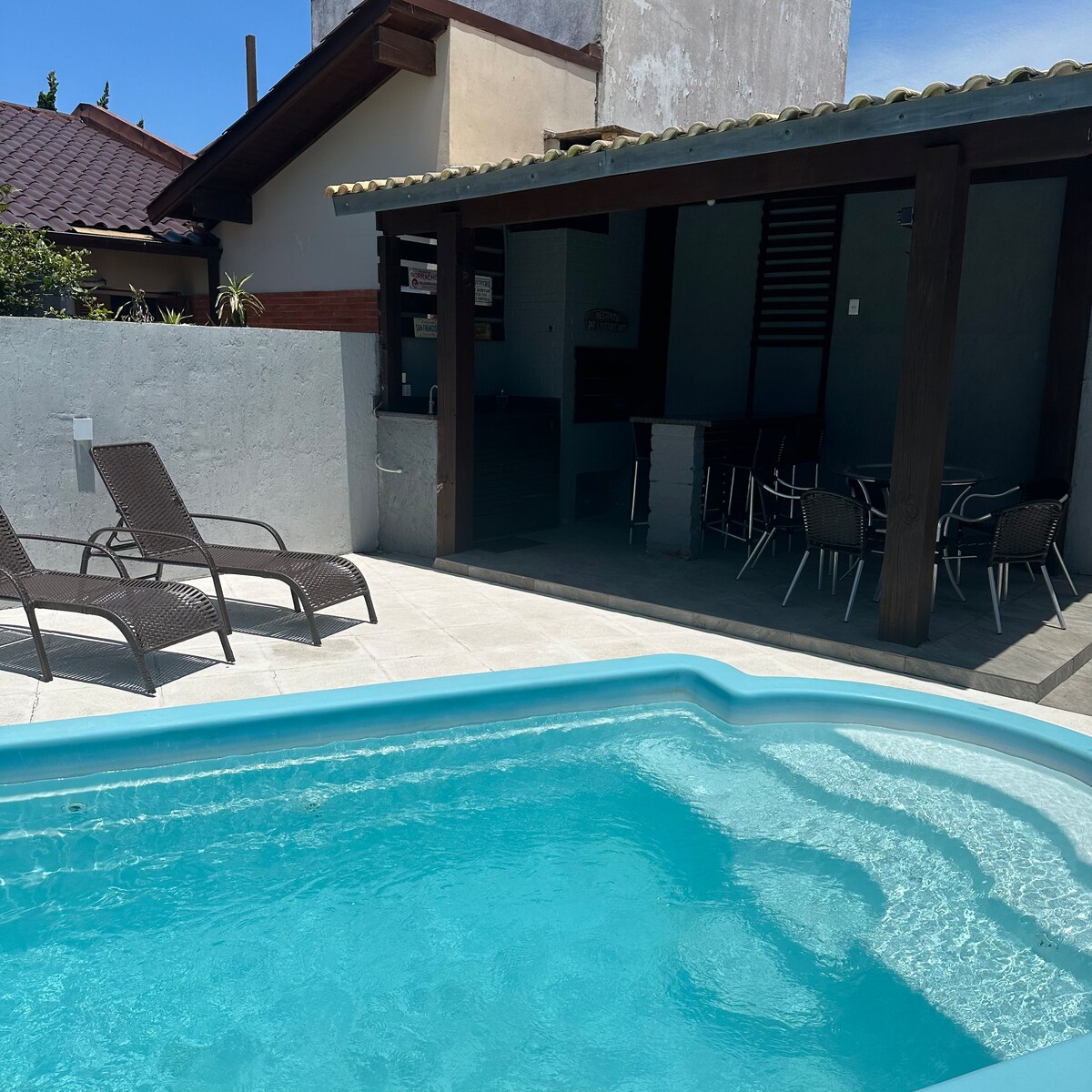 A clear blue pool is visible, surrounded by a tiled area featuring two reclining chairs. An outdoor dining space with a table and multiple chairs is situated beside the pool, sheltered by a roof. The walls are painted in light tones, complementing the sunny environment.