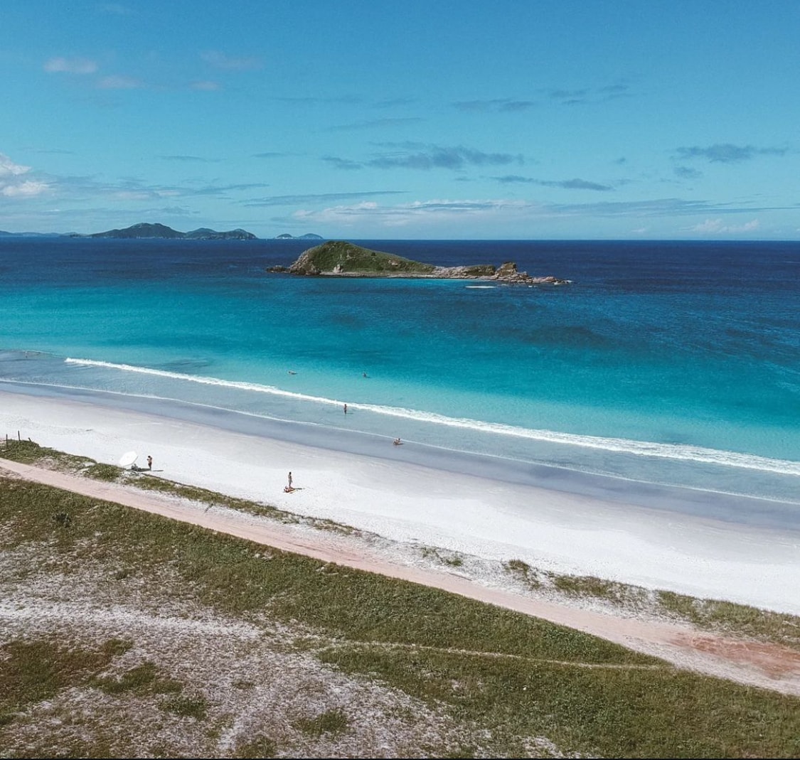 An expansive beach scene features soft white sand meeting crystal-clear blue waters. Gentle waves lap at the shore, while a small island is visible in the distance, surrounded by varying shades of azure. A serene atmosphere is complemented by a clear sky and minimal coastal vegetation.