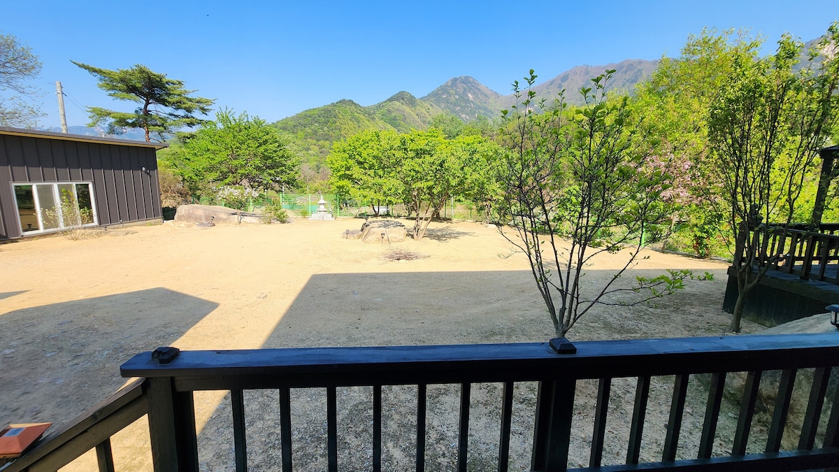 An open outdoor space is surrounded by trees and distant mountains, providing a serene backdrop. The area features a gravel surface and is partially shaded by surrounding structures, allowing for a comfortable environment. Clear skies contribute to the overall bright and welcoming atmosphere.