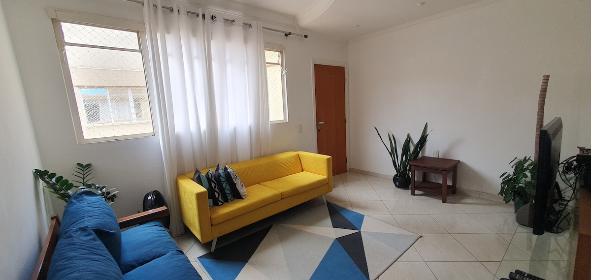 A bright living room features two sofas, one in yellow and another in blue. Light curtains frame the windows, allowing natural light to illuminate the space. A wooden table stands near a potted plant, and a geometric area rug adds a touch of color to the tiled floor.